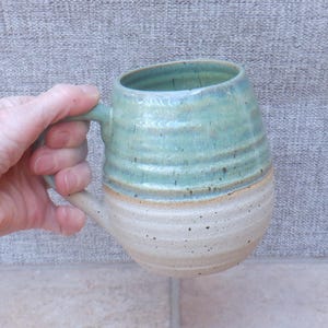 May include: A ceramic mug with a rounded, bulbous shape and a ribbed texture. The upper portion features a glossy, mottled teal and blue glaze, while the lower section is a speckled, matte off-white. It includes a sturdy, curved handle on the side.