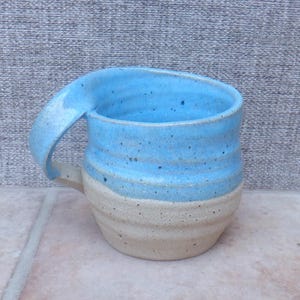 Puede incluir: Una taza de cerámica con diseño bicolor, que presenta una sección superior esmaltada en azul brillante y una base inferior en color beige moteado sin esmaltar. La taza tiene un asa ancha y curva unida al borde superior y estrías horizontales.