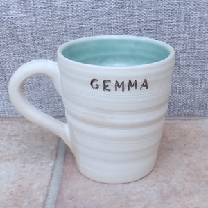 Könnte beinhalten: Weiße Keramiktasse mit hellblauem Innenraum und dem Namen "GEMMA" auf der Seite.