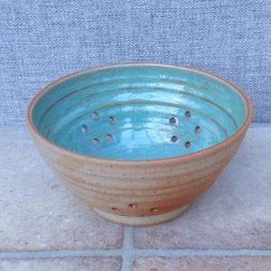 May include: A handmade ceramic colander with a light brown exterior and a turquoise interior. The bowl has a wide rim and numerous drainage holes. The colander is suitable for washing and draining fruits and vegetables.