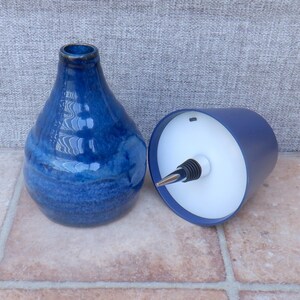 May include: A glossy, deep blue ceramic bottle with a tapered neck and a rounded base, positioned next to a dark blue cylindrical stopper featuring a white interior, a silver-toned metal spout, and a black ribbed rubber seal for a secure fit.