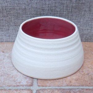 Puede incluir: Tazón de cerámica blanco con interior rojo. El tazón tiene una forma redondeada y un borde ligeramente acampanado.