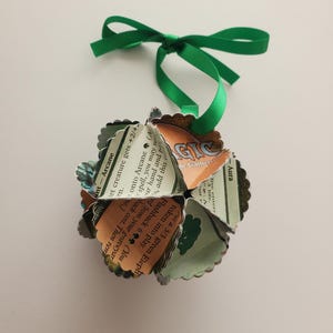May include: A green ribbon tied into a bow hangs from a green and white paper ornament. The ornament is made from folded Magic: The Gathering cards. The text on the cards reads "Ant-Arcane", "Aura", "Flashback 6", "You may return this card from your graveyard to your hand. Then return it to the battlefield. It's flashback cost is equal to its mana cost.", "Put a 3/3 green Elephant token into play.", "Ret creature gets +2/+2.", "When this creature enters the battlefield, you may return target Aura from your graveyard to your hand and put it onto Arcane (A).", "Magic: The Gathering", "De Gather", "3 life."