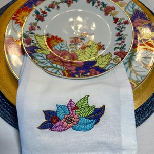Puede incluir: Una servilleta de lino blanco con un dise&ntilde;o floral bordado de colores. La servilleta est&aacute; doblada y colocada encima de un plato con borde dorado y un patr&oacute;n floral.