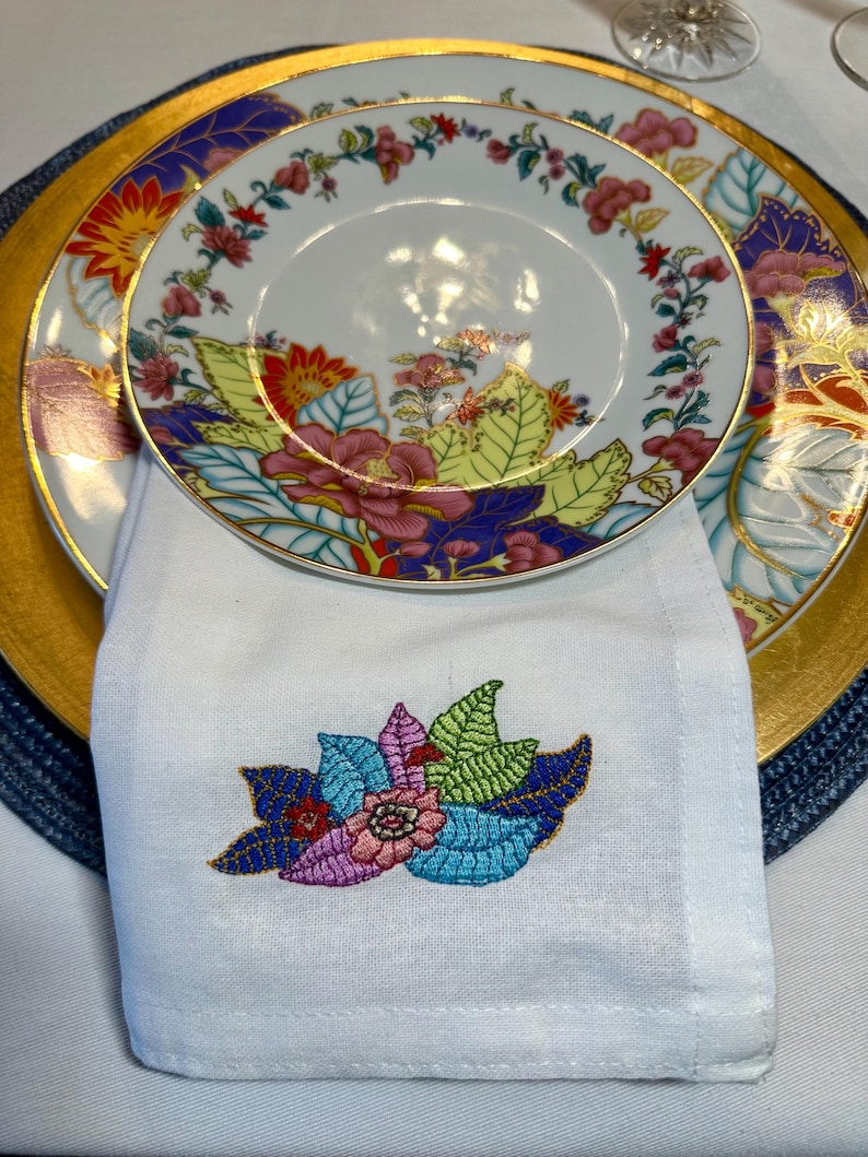 Puede incluir: Una servilleta de lino blanco con un dise&ntilde;o floral bordado de colores. La servilleta est&aacute; doblada y colocada encima de un plato con borde dorado y un patr&oacute;n floral.