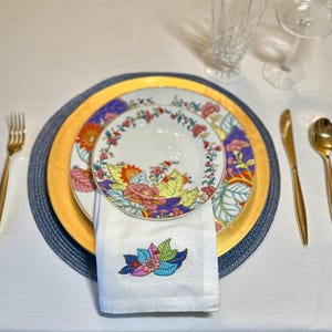 Puede incluir: Una servilleta blanca con un dise&ntilde;o de bordado floral colorido est&aacute; doblada y colocada encima de un juego de tres platos. Los platos est&aacute;n decorados con un patr&oacute;n floral en tonos de rosa, amarillo, rojo y azul. Los platos est&aacute;n sobre un plato base azul y dorado.