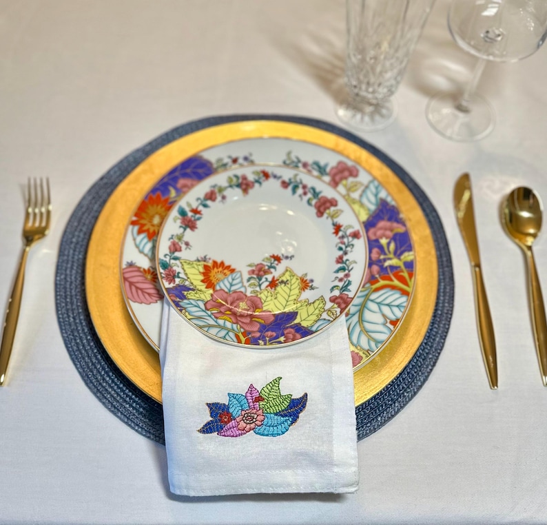 Puede incluir: Una servilleta blanca con un dise&ntilde;o de bordado floral colorido est&aacute; doblada y colocada encima de un juego de tres platos. Los platos est&aacute;n decorados con un patr&oacute;n floral en tonos de rosa, amarillo, rojo y azul. Los platos est&aacute;n sobre un plato base azul y dorado.