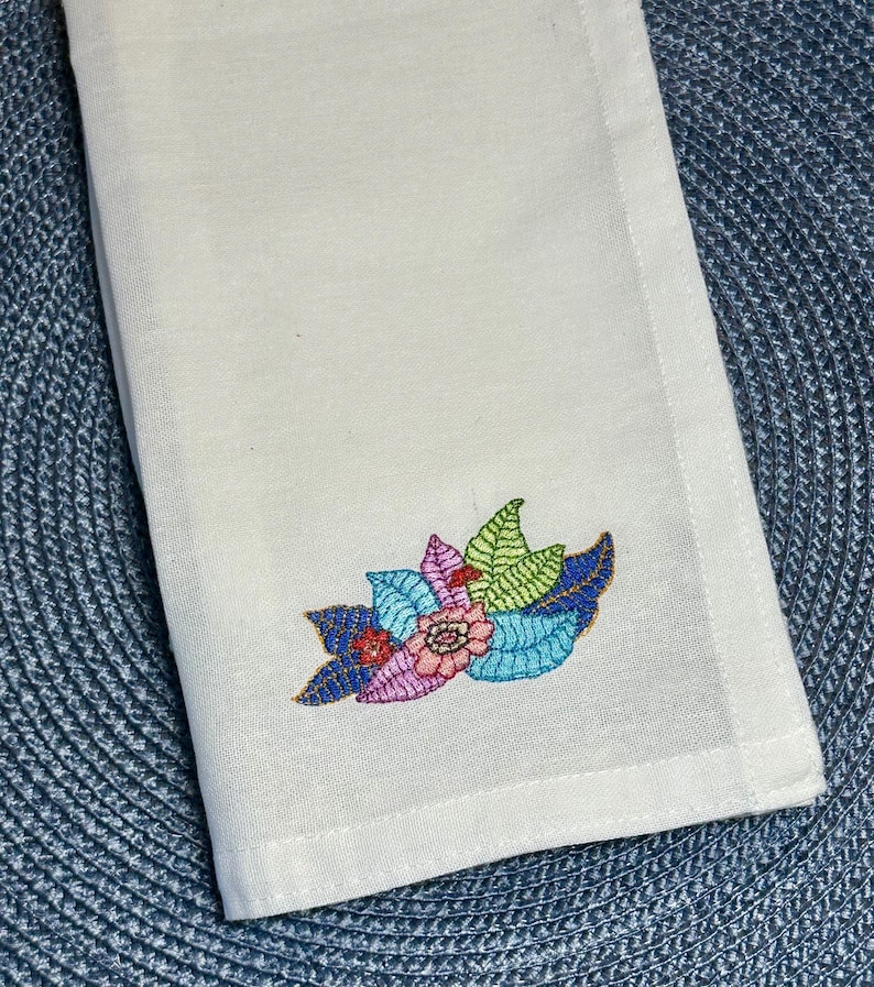 Puede incluir: Servilleta de lino blanco con un dise&ntilde;o bordado de flores y hojas de colores.