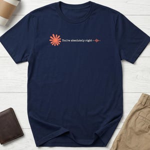 May include: A navy blue short-sleeved t-shirt featuring a graphic on the chest. The design includes an orange flower-like icon, the white text "You're absolutely right", and a small orange sound wave graphic to the right of the text.