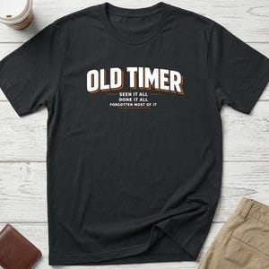 May include: A black short-sleeved t-shirt featuring the white text OLD TIMER in a bold font with an orange outline. Below the main text, smaller white text reads SEEN IT ALL, DONE IT ALL, FORGOTTEN MOST OF IT, separated by horizontal orange lines.