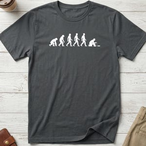 May include: A dark gray short-sleeve t-shirt featuring a white graphic print that depicts the evolutionary progression from a primate to a human, ending with a figure sitting down and interacting with a small electronic device or computer.