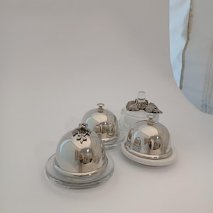 May include: Four decorative butter dishes. Three have silver-coloured, dome-shaped lids with decorative tops, and sit on clear glass or white bases. One dish has a clear glass base and lid with a decorative top.