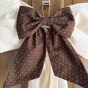 May include: A large brown bow with a floral pattern, hanging against a cream-colored fabric with a similar floral design. The bow is the focal point, with its detailed pattern and contrasting color. The bow is likely a decorative element.
