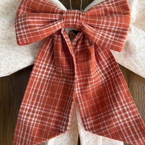 May include: A large, decorative bow featuring a plaid pattern in shades of orange and white. The bow's center is tied, with long, flowing tails. The background is a cream-colored fabric with a subtle floral pattern, creating a layered effect.