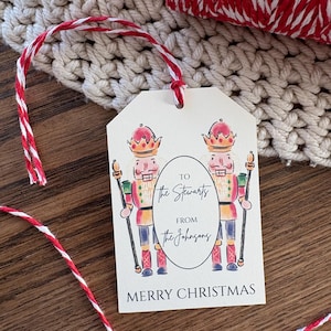 May include: A cream-colored gift tag with a red and white string. The tag features two illustrations of nutcrackers and the text "To the Stewarts from the Johnsons" and "Merry Christmas".