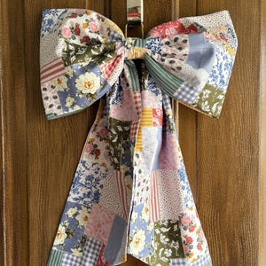 May include: A large, decorative bow made from patchwork fabric, featuring floral and geometric patterns in shades of pink, blue, green, and red. The bow is suspended from a metal hook, creating a charming, handcrafted accent piece.