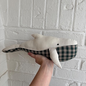 May include: A plush whale toy with a cream-colored body and a green and black plaid tail. The whale has a small black eye and a white fin. The toy is held in front of a white brick wall.