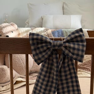 May include: A large, plaid bow in navy blue and tan hangs on a wooden crib. The bow is the focal point, with its symmetrical design and long, flowing tails. The crib is filled with soft bedding and a teddy bear.