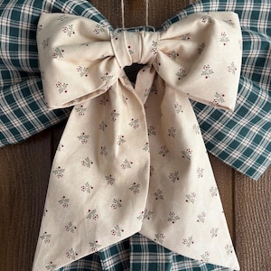 May include: A large decorative bow with a cream-colored center and green plaid wings. The cream-colored fabric is patterned with small red and green floral sprigs. The bow is likely used for seasonal or holiday decor.