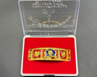 May include: A gold-colored ring with red and blue enamel accents, displayed in a clear plastic case with a red interior. The ring features intricate designs and a central figure.