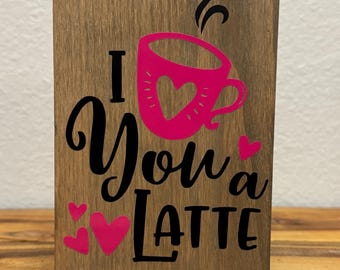 Peut inclure: Une pancarte en bois avec le texte "I You a Latte" en lettres noires. La pancarte présente une tasse de café rose avec un cœur et de la vapeur qui sort du haut. Il y a aussi des cœurs roses autour du texte.
