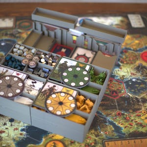 Può includere: Un organizer per giochi da tavolo in plastica grigia con scomparti multipli contenenti vari componenti di gioco, tra cui quadranti circolari, piccoli gettoni in legno e carte, posizionato sopra un grande tabellone di gioco illustrato.