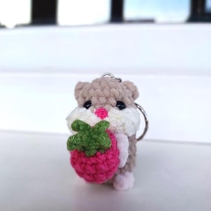 May include: A crocheted hamster keychain holding a pink and green strawberry. The hamster is beige with white fur around its face and has black eyes.
