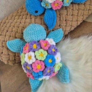May include: Two crocheted blue turtle toys with colourful flower accents. The turtles are on a brown and white fluffy surface.