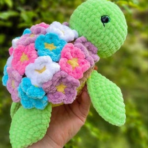 May include: A crocheted green turtle with a shell made of colorful flowers. The turtle is holding a flower in its mouth.