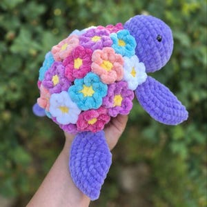 May include: A plush, purple turtle with a shell covered in colorful, flower-shaped accents. The flowers are in shades of pink, blue, peach, and white, with yellow centers. The turtle's head and limbs are a solid purple color.
