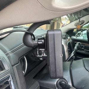 May include: Black electronic device mounted on a vehicle's dashboard, near the steering wheel. The device is rectangular with a small button and is connected to a black cable. The dashboard is black with white stitching.