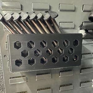 May include: A black metal magazine holder with a honeycomb pattern. The holder is mounted on a gray pegboard wall and contains a stack of silver metal magazines.