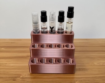 May include: A rose gold 3-tiered perfume bottle organizer with 15 slots. The organizer is made of plastic and has a textured surface. There are five perfume bottles in the top row of the organizer.