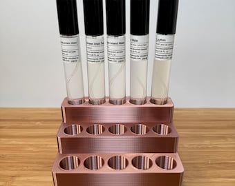 May include: A rose gold metal stand with three tiers, each tier has six round holes to hold small bottles. Five clear glass bottles with black caps are displayed on the top tier. The bottles are labeled with the names of different fragrances: Mountain Water, Green Irish Tweed, High Island Water, Le Male, and Layton.