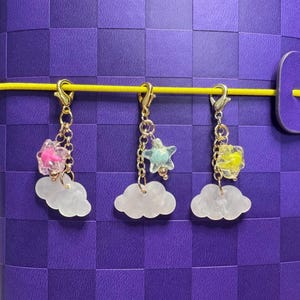May include: Three charm keyrings with gold-coloured clasps and chains. Each keyring features a white cloud charm, topped with a star or flower-shaped charm in pink, blue, or yellow. The charms hang against a purple background.