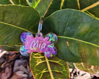 Puede incluir: Una placa de identificación para perro en forma de hueso rosa y morado con purpurina y flores prensadas. La placa tiene el nombre "Mia" grabado en ella.