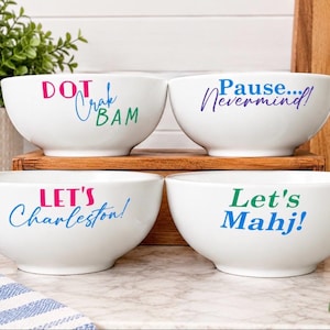 May include: Four white ceramic bowls featuring colorful text. The top left bowl reads DOT Crak BAM. The top right bowl reads Pause... Nevermind!. The bottom left bowl reads LET'S Charleston!. The bottom right bowl reads Let's Mahj! in blue and green.