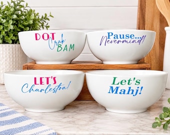 May include: Four white ceramic bowls featuring colorful text. The top left bowl reads DOT Crak BAM. The top right bowl reads Pause... Nevermind!. The bottom left bowl reads LET'S Charleston!. The bottom right bowl reads Let's Mahj! in blue and green.