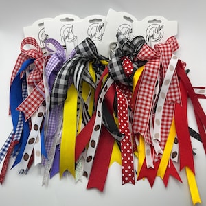 May include: A collection of colorful ribbon hair accessories with various patterns, including gingham, polka dots, and coffee bean designs. Each accessory features a bow and multiple streamers in red, blue, yellow, black, and white. The accessories are attached to white cardstock.