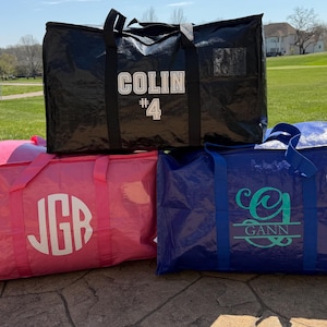 May include: Three large, personalized storage bags in black, pink, and blue. The black bag has "COLIN #4" in white. The pink bag has "JGB" in white. The blue bag has "GANN" in teal.