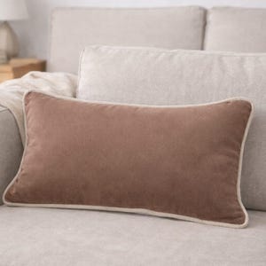 Può includere: Un cuscino rettangolare in velluto marrone con un bordo color crema è appoggiato su un divano beige chiaro. Il cuscino offre un contrasto caldo con i toni neutri del divano e dell'arredamento circostante.