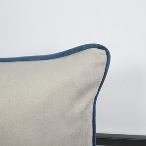May include: Close-up of a square pillow with a light gray fabric and a dark blue trim. The pillow has a soft, textured appearance, and the trim neatly outlines the edges. The design is simple and elegant, suitable for home decor.