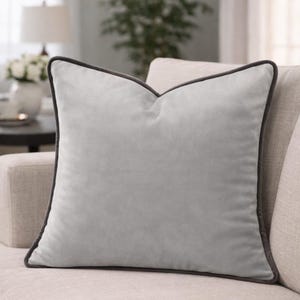 Puede incluir: Un cojín cuadrado de terciopelo gris claro con un ribete gris oscuro. El cojín está sobre un sofá de color claro. Es un elemento decorativo para una sala de estar o un dormitorio.