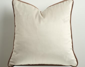 May include: A square, cream-colored pillow with a brown border. The pillow is made of a soft, velvety material and is placed on a white surface. The pillow has a simple, elegant design.
