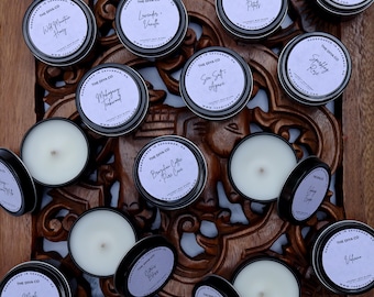 May include: A collection of round metal candle tins with white labels featuring the text The Diya Co, handpoured in Savannah, and various scent names. The candles contain white wax with visible wicks, some with lids removed to show the wax surface.