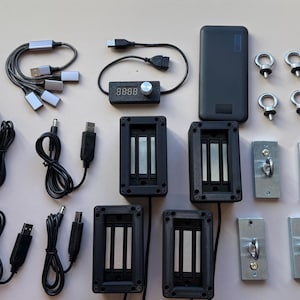 May include: An assortment of electronic components, including a power bank, cables, and magnetic blocks, arranged on a light surface. The items are primarily black and silver, with a digital display and metal eyelets.