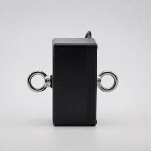 May include: A black rectangular object with two silver-colored, circular eye bolts attached to its sides. A small, black wire extends from the top. The object is set against a plain, light background.