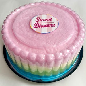 May include: A multi-colored cotton candy cake in a clear plastic container with a pink lid. The lid has a sticker that says "Sweet Dreamz" in white letters with a pink and white striped background.