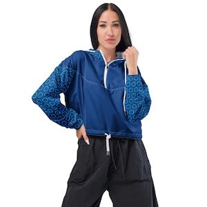 May include: A blue cropped windbreaker jacket with a white zipper and drawstring. The sleeves feature a blue and teal circular pattern. The jacket is worn with black pants.