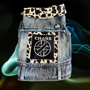 May include: A denim dog vest with a cow print collar and back patch. The back patch features the word "CRASS" above a white graphic. A small "Punk Pet" pin is on the back.